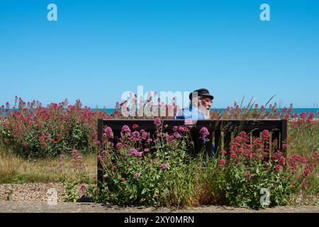 Alter Mann mit Fischerhut, der auf einer Bank am Strand in Aldeburgh, Suffolk, England sitzt. Stockfoto
