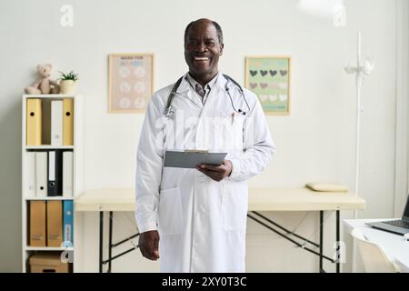 Afroamerikaner Arzt steht in der medizinischen Praxis, trägt weißen Mantel und Stethoskop und hält das Klemmbrett, während er lächelt. Hintergrund: Bürobedarf und dekorative Poster Stockfoto