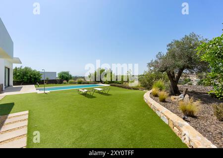 Eine moderne weiße Villa mit einem begrünten Hinterhof mit Kunstrasen, einem Olivenbaum und einem privaten Swimmingpool unter der blauen Ske Stockfoto