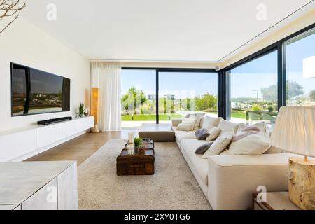 Ein luxuriöses, modernes Wohnzimmer verfügt über ein großes weißes Sofa und große Fenster, die eine üppige grüne Landschaft freigeben Stockfoto