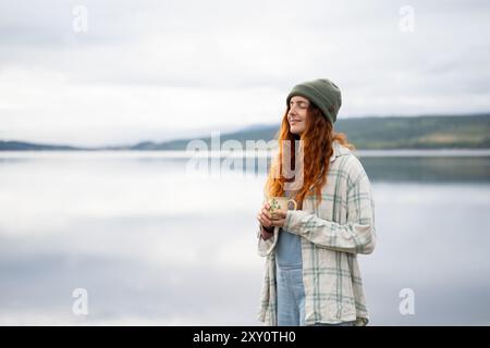 Eine junge Frau mit rotem Haar, die eine gemütliche Mütze und ein kariertes Hemd trägt, genießt einen friedlichen Moment bei einer Kaffeetasse am ruhigen See Stockfoto