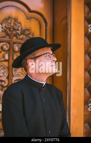 Ein katholischer Priester in traditioneller schwarzer Kleidung blickt nachdenklich nach oben und steht vor den verzierten Holztüren einer orthodoxen katholischen Kirche in Al Stockfoto