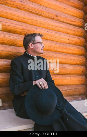 Katholischer Priester in kontemplativer Pose, draußen vor rustikaler Holzkulisse. Er schaut zur Seite, nicht direkt auf die Kamera und hält Stockfoto