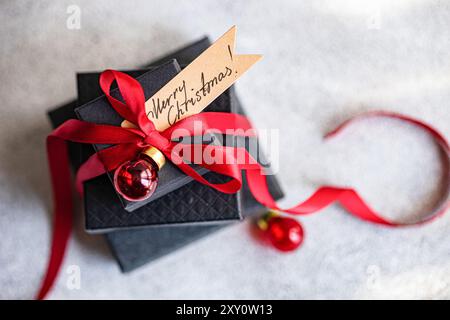 Blick von oben auf schwarze Geschenkboxen, die elegant mit einem roten Band gebunden und mit einem roten Weihnachtsschmuck verziert sind. Auf der beigefügten Grußkarte steht Merry Christma Stockfoto