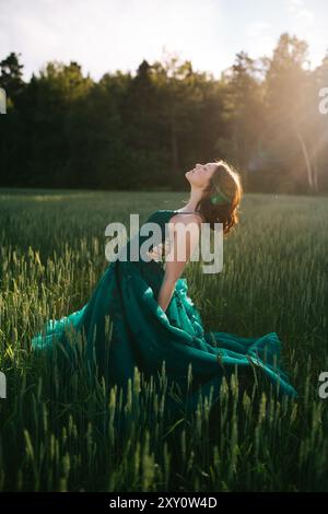 Ein Mädchen im Teenageralter in einem leuchtend blauen Ballkleid schwelgt bei Sonnenuntergang in einem üppigen grünen Feld, wobei das Sonnenlicht durch die Bäume im Hintergrund gefiltert wird Stockfoto