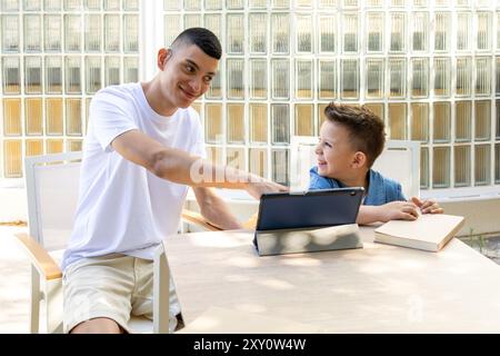 Ein junger Mann mit Zerebralparese und leichter Taubheit macht sich mit einem kleinen Jungen in einer Lerneinheit mit einem Tablet beschäftigt. Er sitzt an einem Tisch im Freien, gemeinschaftlich Stockfoto