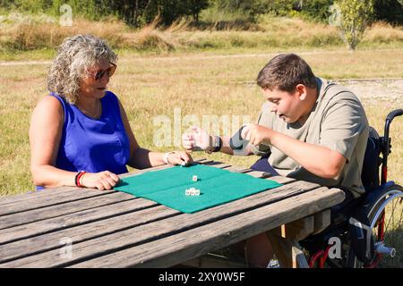 Ein kleiner Junge mit Zerebralparese, sitzt im Rollstuhl und spielt aktiv mit einer lächelnden Frau an einem Holztisch im Freien, exempli Stockfoto