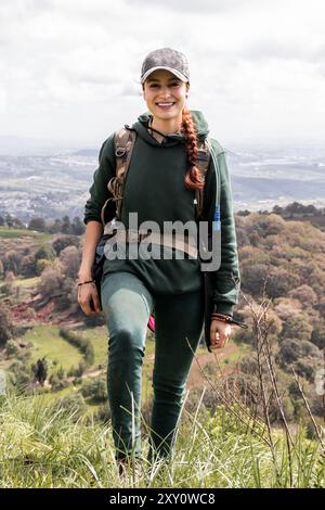 Eine junge Kaukasierin, die beim Wandern in der Natur für die Kamera lächelt, in Outdoor-Ausrüstung gekleidet und einen Rucksack trägt, genießt ein atemberaubendes Tal Stockfoto