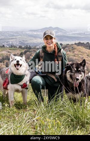 Lächelnde Wanderer mit zwei Huskyhunden auf einem Bergweg. Sie trägt Wanderausrüstung und die Hunde tragen rote Westen. Malerische Aussicht auf die Roller Stockfoto