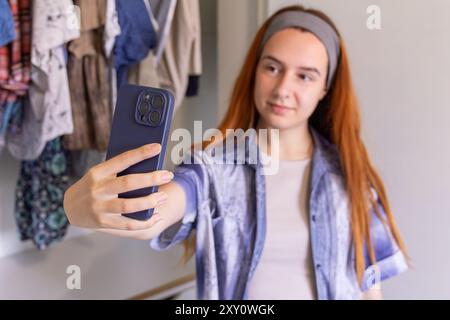 Eine junge Frau mit langen roten Haaren, die ein Hemd trägt, organisiert ihre Garderobe. Sie schaut in die Kamera, während sie ihr Smartphone hält, um eine Abkürzung zu machen Stockfoto