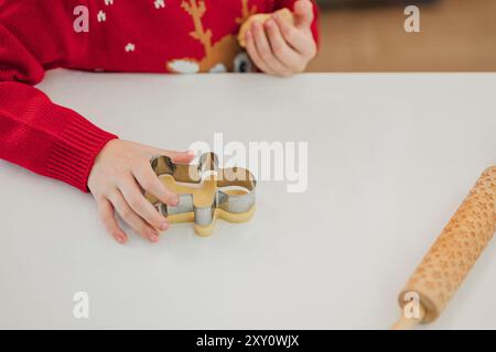 Eine Nahaufnahme eines Kindes, eingewickelt in rote Weihnachtspullover, die für die Feiertage backen. Ein Kind drückt einen Lebkuchenkeksausstecher in den doug Stockfoto