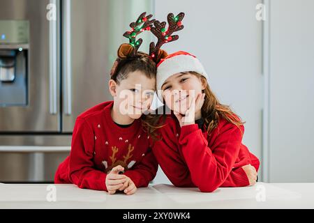 Fröhliche Zwillingsgeschwister in festlichen Weihnachtspullover gekleidet, einer trägt Rentiergeweih und der andere einen Weihnachtsmann-Hut, lächeln fröhlich, während sie in A posieren Stockfoto