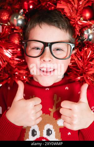 Ein fröhlicher kleiner Junge, der eine schwarze Brille und einen roten Weihnachtspullover mit Rentier trägt, gibt die Daumen hoch, während er von einem leuchtend roten festlichen d umgeben ist Stockfoto