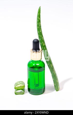 Ein lebhaftes Bild, das eine grüne Tropfflasche aus Glas mit Aloe Vera-Essenz sowie ein frisches Aloe Vera-Blatt und geschnittene Segmente zeigt, isoliert auf einem weißen Stockfoto
