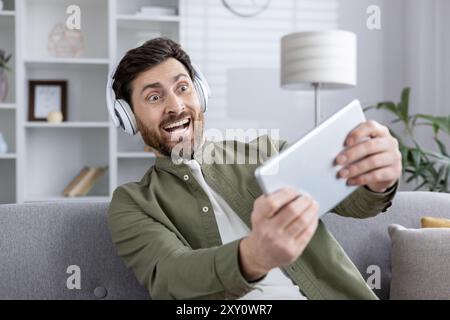 Fröhlicher Mann mit Kopfhörern, der das Tablet hält und sich intensiv auf Videospiele konzentriert. Drückt Begeisterung und Engagement für modernes Gaming-Erlebnis mit Telefon und Technologie aus. Stockfoto