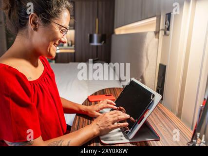 Eine fröhliche Frau in rotem Kleid arbeitet mit einem digitalen Tablet in einem elegant eingerichteten Hotelzimmer und unterstreicht die Integration von Technologie Stockfoto