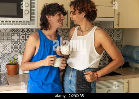 Ein schwules Paar teilt sich einen freudigen Moment, während es in seiner gemütlichen Küche Kaffeetassen hält und Wärme und Gesellschaft zum Ausdruck bringt Stockfoto