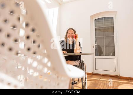 Eine freudige Frau im Rollstuhl führt Geschäfte von zu Hause aus, nutzt ihr Telefon und spricht sich in einem hell beleuchteten Raum herzlich mit einem Kunden an Stockfoto