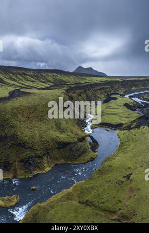 Ein ruhiger Blick aus der Luft auf das isländische Hochland mit üppigem Grün und einem sich windenden Fluss, der sich durch bergiges Gelände unter einem stimmungsvollen Himmel schlängelt. Stockfoto