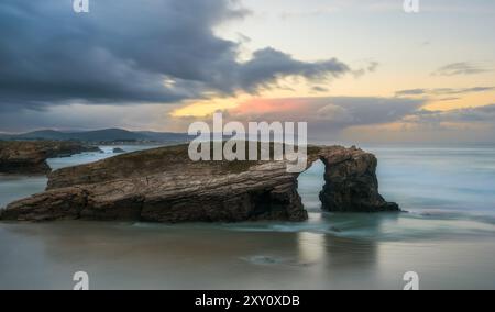 Eine ruhige Küstenlandschaft mit einem natürlichen Felsbogen, beleuchtet von einem sanften Sonnenuntergang vor einem dramatischen Himmel. Stockfoto