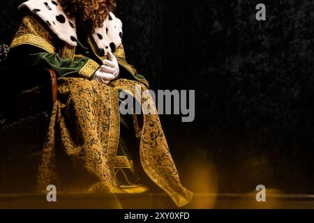 Einer der drei Weisen, Gaspar, in einem extravaganten königlichen Anzug, der mit Goldstickerei und Pelzen verziert ist, sitzt majestätisch in einer dunklen Umgebung Stockfoto