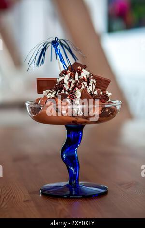 Ein luxuriöser Schokoladeneis-Eisbecher mit Schlagsahne, reichhaltiger Schokoladensoße, Streuseln und Schokoladenstückchen, serviert in einem stilvollen blauen gla Stockfoto