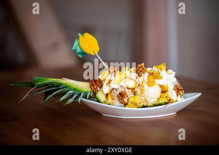 Leckeres Ananasboot-Dessert mit Vanilleeis garniert mit Walnüssen, Ananaswürfeln und Popcorn auf einer weißen Keramikplatte. Stockfoto