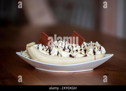 Ein köstlicher Bananensplitt mit Schlagsahne, Schokoladensirup, Streuseln und Schokoladenstückchen, serviert in einer weißen Schüssel auf einem Holztisch. Stockfoto
