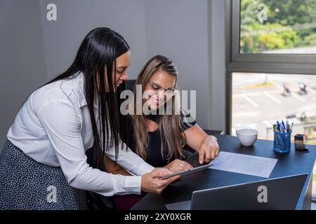 Zwei lateinamerikanische Anwältinnen, die sich ein digitales Tablet anschauen, während sie einen Fall in einem modernen Büro diskutieren. Sie sitzen an einem Schreibtisch mit einem Lapt Stockfoto