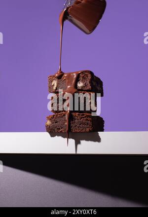 Stapel hausgemachter Schokoladen-Brownies, die reich mit Nüssen gefüllt sind und großzügig mit einer glänzenden Schokoladensauce gegen ein markantes Violett getränkt werden Stockfoto