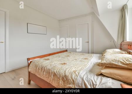 Ein einfaches und einladendes Schlafzimmer im Dachgeschoss mit einem komfortablen Bett mit beigefarbener Bettwäsche, gerahmten Kunstwerken und Tageslicht. Stockfoto