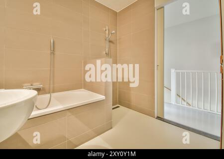 Ein elegantes, modernes, minimalistisches beigefarbenes Badezimmer mit weißer Badewanne, Duschvorrichtung, Waschbecken und Sicherheitsgeländer in der Nähe der Glastür. Stockfoto