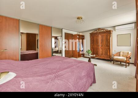 Ein Schlafzimmer im Retro-Stil mit großem Bett mit malvenfarbenen Bezügen, Holzschrank, klassischem Sessel und Spiegelschranktüren. Stockfoto