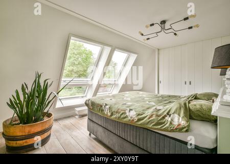 Modernes Schlafzimmer im Dachgeschoss mit großen Dachfenstern, einem komfortablen Bett mit einzigartiger Tagesdecke und einer großen Topfpflanze. Stockfoto