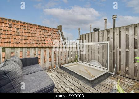 Eine einladende, urbane Dachterrasse mit Holzterrasse, Dachfenster, gemütlichem Sofa im Freien und rustikalem Zaun unter blauem Himmel. Stockfoto