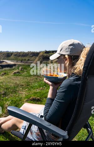 Eine junge Frau sitzt bequem vor ihrem Wohnmobil und genießt ein sonniges Mittagessen inmitten üppiger Natur Stockfoto