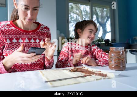 Mutter schaut sich ein Rezept für Weihnachtssüße auf ihrem Handy an, das sie mit ihrem Sohn in der Küche ihres Hauses in Weihnachtskleidung kochen kann Stockfoto