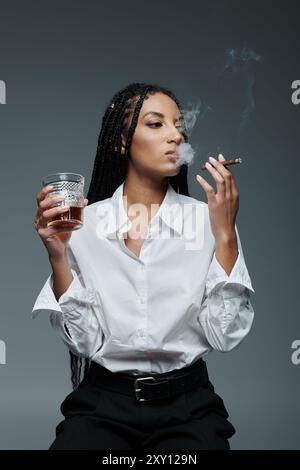 Eine junge Frau in einem weißen Hemd und einer schwarzen Hose raucht eine Zigarette und hält ein Glas Alkohol. Stockfoto