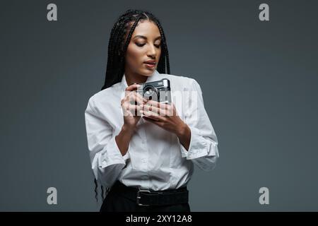 Eine junge Frau in einem weißen Hemd steht vor einem grauen Hintergrund, hält eine Kamera in der Hand und fokussiert auf das Objektiv. Stockfoto