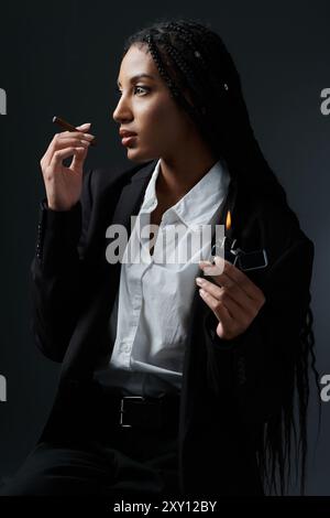 Eine junge Frau in weißem Hemd und schwarzem Blazer posiert vor grauem Hintergrund, hält ein Feuerzeug und eine Zigarette in der Hand. Stockfoto