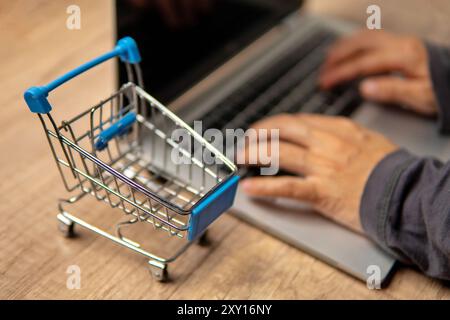 Nahaufnahme des Mini-Einkaufswagens und der Hände, die auf dem Laptop tippen. Online-Shopping-Konzept. Stockfoto
