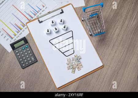 Eine Zwischenablage mit einem Verkaufstrichdiagramm und Würfeln, die Kunden repräsentieren. Geld und ein Einkaufswagen sind in der Nähe, was Umsätze und Umsätze symbolisiert. Stockfoto