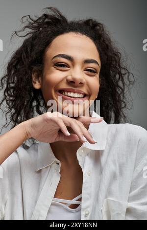 Eine junge Frau mit lockigen Haaren lächelt hell, während sie vor grauem Hintergrund posiert. Stockfoto