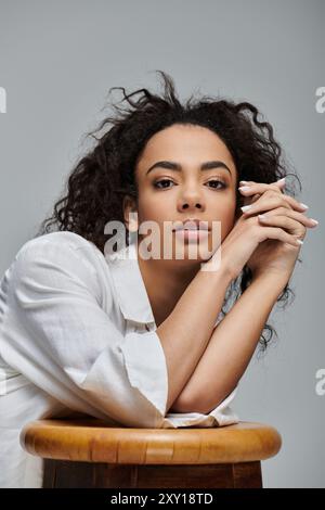 Eine schöne junge Frau mit dunklen lockigen Haaren beugt sich nach vorne und legt ihre Arme auf einen hölzernen Hocker vor grauem Hintergrund. Stockfoto