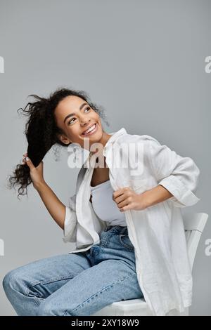 Eine junge schwarze Frau sitzt auf einem weißen Stuhl vor grauem Hintergrund und wirft spielerisch ihre Haare, während sie vor Freude strahlt. Stockfoto