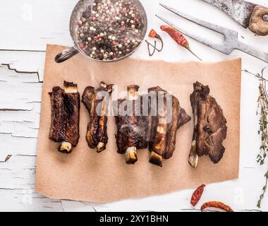 Blick von oben auf gegrillte Schweinerippchen mit Kräutern auf dem Stück braunem Papier auf weißem Holzhintergrund mit aufgerissener Farbe. Draufsicht Stockfoto