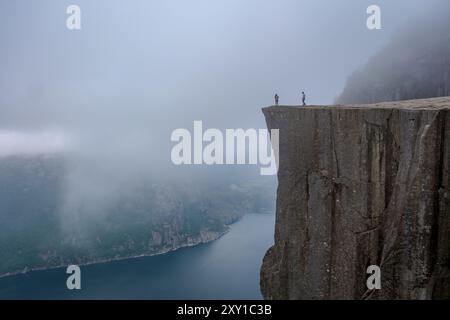 Zwei Abenteurer stehen auf einer zerklüfteten Klippe und beobachten den wirbelnden Nebel über einem atemberaubenden norwegischen Fjord. Die ätherische Landschaft besticht durch ihre natürliche Schönheit. Preikestolen Norwegen Stockfoto