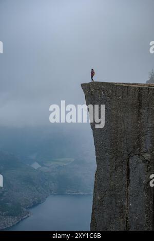 Ein einsamer Wanderer steht triumphierend an einer Klippe und blickt in den dramatischen, nebelumhüllten Fjord, der in die atemberaubende Schönheit der Natur eingetaucht ist. Preikestolen Norwegen Stockfoto