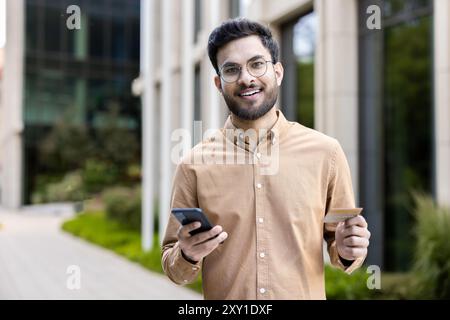 Lächelnder junger Mann mit Smartphone und Kreditkarte, Online-Shopping und digitalem Bezahlen. Person, die praktische mobile Transaktionen in einem urbanen Außenbereich genießt. Stockfoto