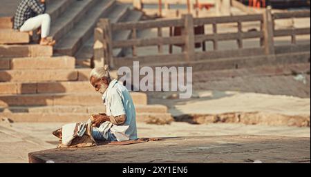 Varanasi, Indien. Der Alte Bettler Zählt Seine Einnahmen. Ein Alter Mann Sitzt Am Ganges Embankment. Der Senior-Mann Zählt Sein Geld. Vormittag Im Heiligen Varanasi Ghats Stockfoto